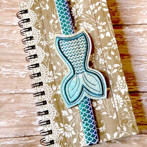 Puede incluir: Un marcap&aacute;ginas de cola de sirena azul y verde con detalles de purpurina en una libreta con estampado marr&oacute;n y blanco.