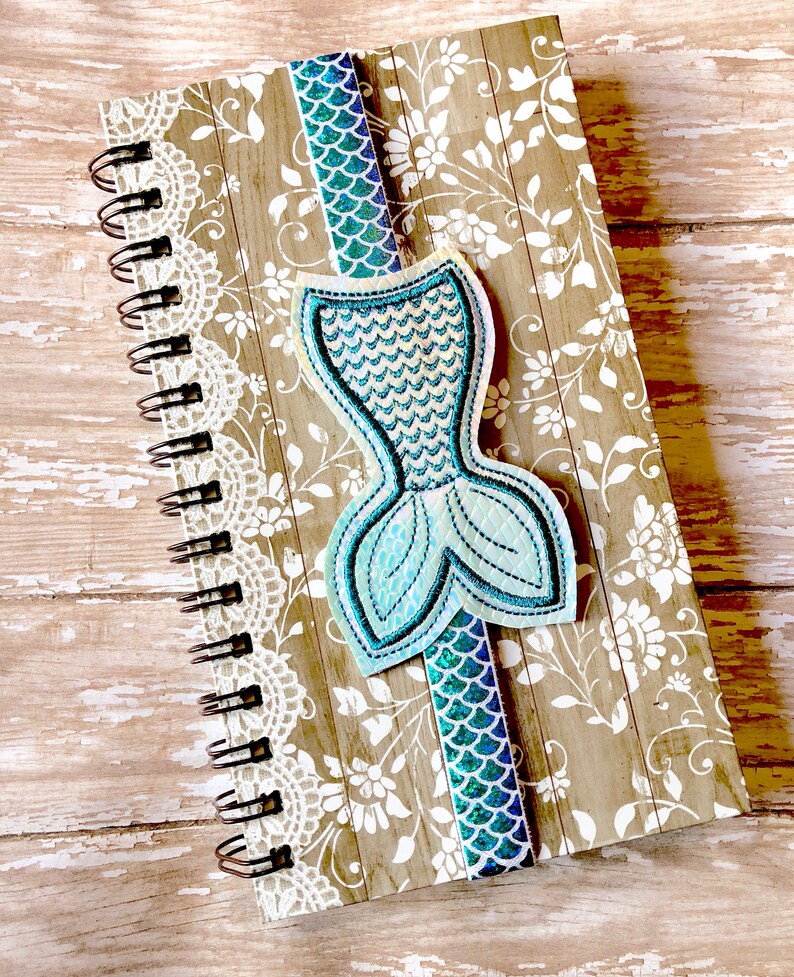 Puede incluir: Un marcap&aacute;ginas de cola de sirena azul y verde con detalles de purpurina en una libreta con estampado marr&oacute;n y blanco.
