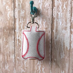 May include: A white glitter hand sanitiser holder shaped like a baseball with red stitching. The holder is attached to a silver key chain and is hanging on a teal hook.