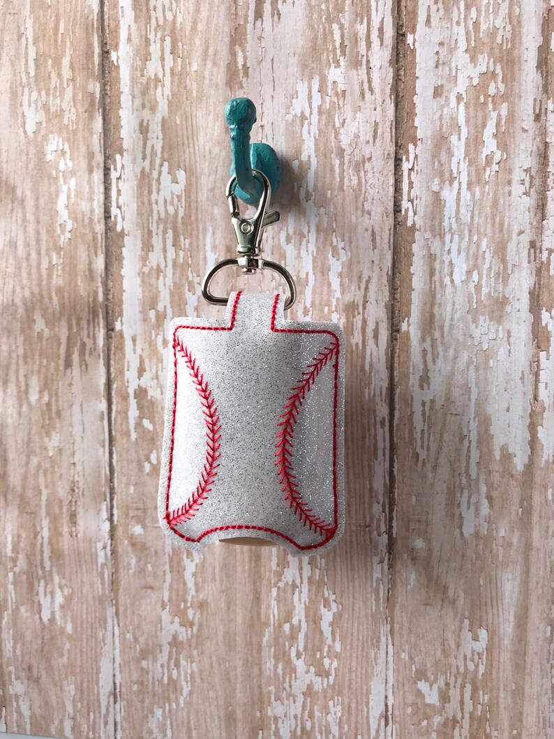 May include: A white glitter hand sanitiser holder shaped like a baseball with red stitching. The holder is attached to a silver key chain and is hanging on a teal hook.