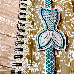 Puede incluir: Un marcap&aacute;ginas con forma de cola de sirena con escamas iridiscentes en tonos azules y verdes. El marcap&aacute;ginas est&aacute; unido a un cuaderno de espiral con un dise&ntilde;o floral.