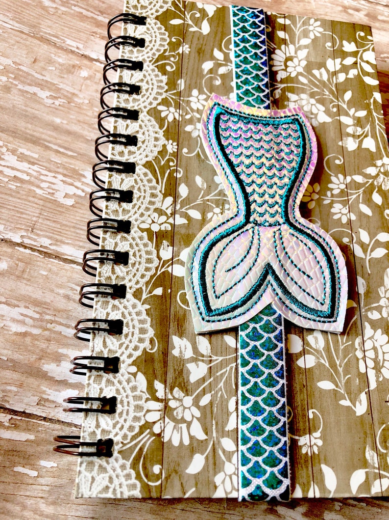 Puede incluir: Un marcap&aacute;ginas con forma de cola de sirena con escamas iridiscentes en tonos azules y verdes. El marcap&aacute;ginas est&aacute; unido a un cuaderno de espiral con un dise&ntilde;o floral.