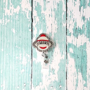 May include: A white and grey sock monkey face with a red hat, attached to a clear retractable badge holder.