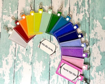 May include: A rainbow assortment of 10 keychains with a white glitter finish. Each keychain has a white button and a silver ring. The keychains are arranged in a semi-circle on a light blue wooden surface. The text "while ellie dreams" is printed on two white cards.