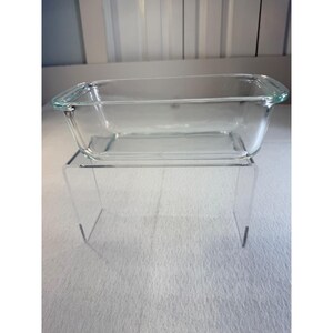 May include: A clear rectangular glass loaf pan with integrated handles on both ends. The bakeware features smooth, transparent walls and a flat base, designed for baking breads or cakes, showcasing a simple, functional, and durable construction.