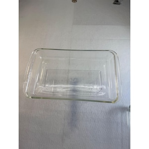 May include: A clear rectangular glass baking dish with rounded corners and integrated handles on the short sides. The center of the base features a circular embossed logo with text that is partially visible through the transparent glass material.
