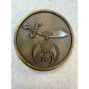 May include: A circular bronze-toned metal medallion featuring a textured background. The design displays a horizontal scimitar sword positioned above a crescent moon emblem, which incorporates a central pharaonic face and a five-pointed star.