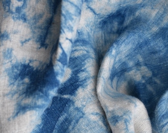 Puede incluir: Una tela ligera con un patrón de teñido anudado en colores azul intenso y blanco. El material tiene una textura tejida visible con pliegues y arrugas que crean profundidad, mostrando una mezcla de formas abstractas en tono índigo sobre una base gris.