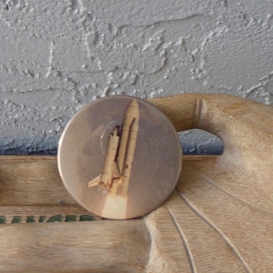 May include: A circular pinback button featuring a graphic illustration of a space shuttle during launch. The design displays the white and orange shuttle against a light background, with a plume of smoke visible at the base of the rocket.