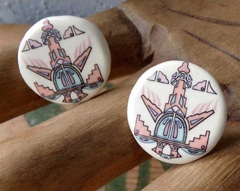 May include: A pair of white ceramic earrings with a geometric design in pink, blue, and black. The design features a stylized building with a pointed roof and a series of windows and doors.