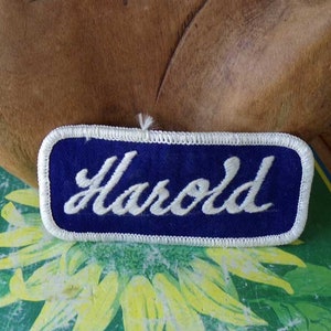 May include: A blue felt name tag with white stitching and the name "Harold" embroidered in white.