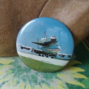 May include: A round button with a blue sky background and a white space shuttle on top of a white airplane. The shuttle and airplane are on a runway with green grass.