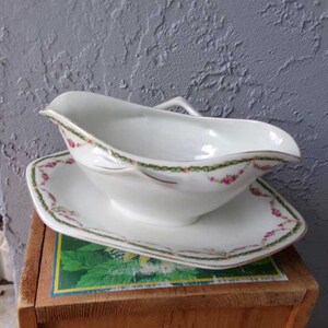 May include: White porcelain gravy boat with a green and pink floral design. The boat has a handle and sits on a matching plate.