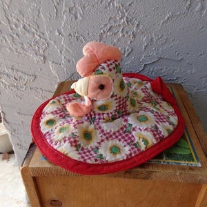 May include: A red, white, and yellow quilted hot pad shaped like a chicken. The chicken has a pink comb and wattle, black eyes, and a white beak.