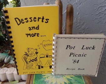 May include: Two vintage recipe books with yellow and white covers. The yellow book has a cartoon illustration of a bear and the text "Desserts and more... Pot Luck Picnic 1985". The white book has the text "Pot Luck Picnic '84 Recipe Book".