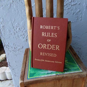 May include: A red hardcover book titled "Robert's Rules of Order Revised" with gold lettering. The book is the 75th Anniversary Edition.