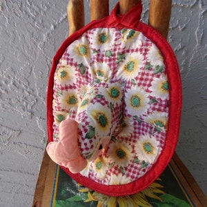 May include: Red and white pot holder with a sunflower pattern and a pink chicken head. The pot holder is made of fabric and has a red trim.