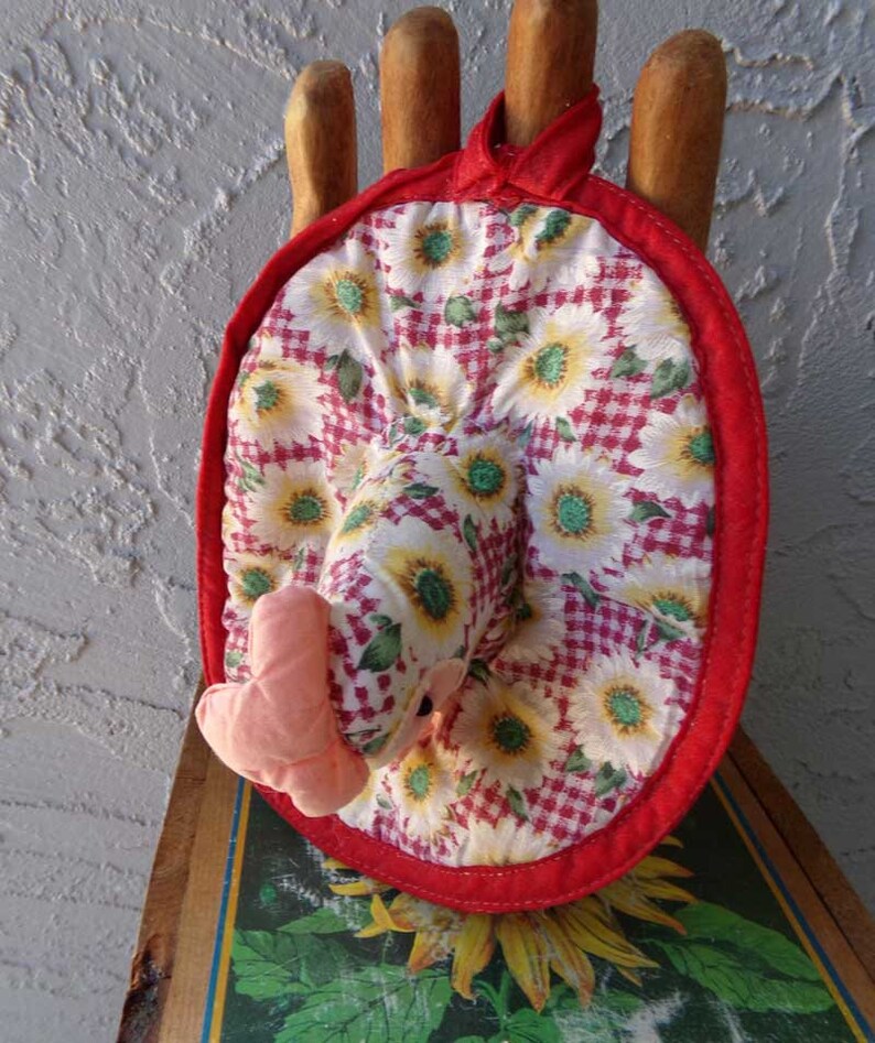 May include: Red and white pot holder with a sunflower pattern and a pink chicken head. The pot holder is made of fabric and has a red trim.