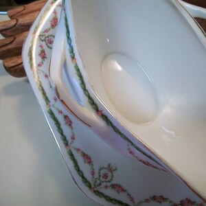 May include: A white porcelain gravy boat with a floral design in pink and green. The boat has a handle on each side and a spout on one end.