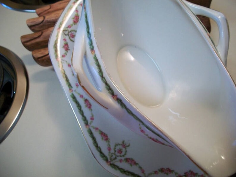 May include: A white porcelain gravy boat with a floral design in pink and green. The boat has a handle on each side and a spout on one end.