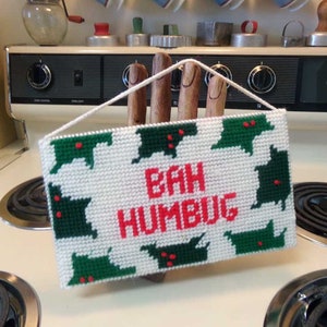 May include: A white, rectangular, needlepoint sign with the words "BAH HUMBUG" in red letters. The sign is decorated with green Christmas tree shapes with red accents. A white string is attached for hanging.