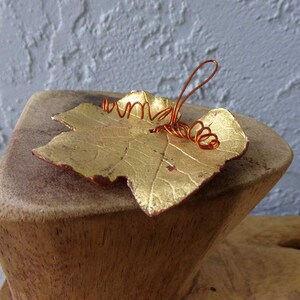May include: A gold-colored leaf with a copper wire stem and tendrils. The leaf has a textured surface and is resting on a wooden block. The leaf's edges are a reddish-brown color.