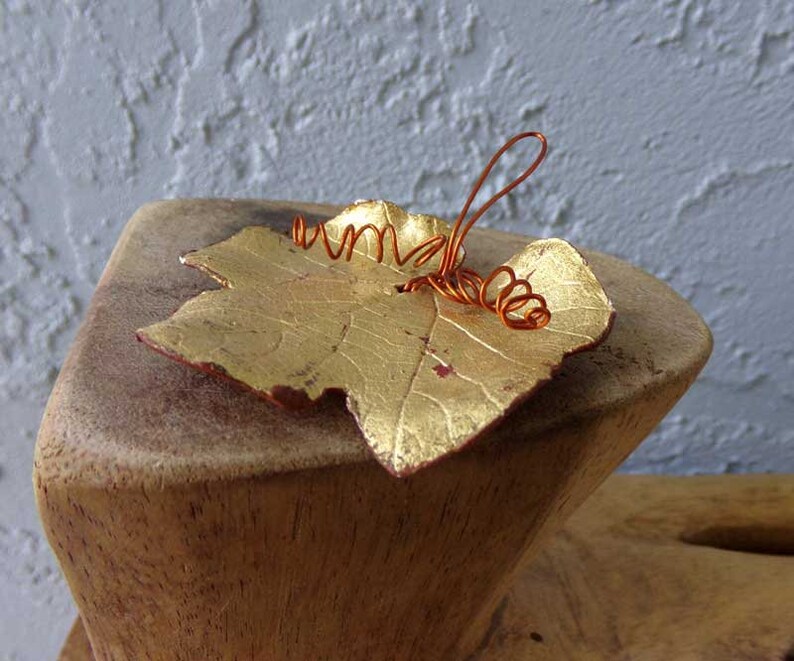 May include: A gold-colored leaf with a copper wire stem and tendrils. The leaf has a textured surface and is resting on a wooden block. The leaf's edges are a reddish-brown color.