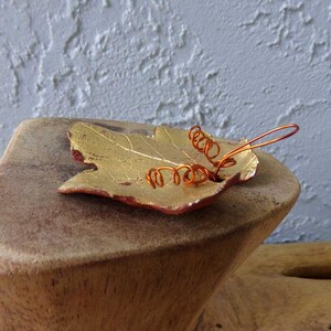 May include: A handcrafted leaf-shaped dish with a gold finish and red edges. Copper wire spirals are attached to the leaf. The dish is displayed on a wooden stand, creating a rustic and artistic aesthetic.