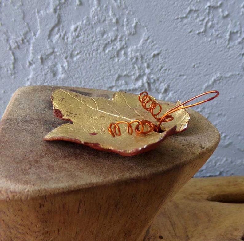 May include: A handcrafted leaf-shaped dish with a gold finish and red edges. Copper wire spirals are attached to the leaf. The dish is displayed on a wooden stand, creating a rustic and artistic aesthetic.