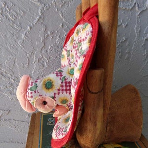 May include: Red and white quilted oven mitt shaped like a chicken with a pink head and black eyes. The mitt is hanging on a wooden hand.