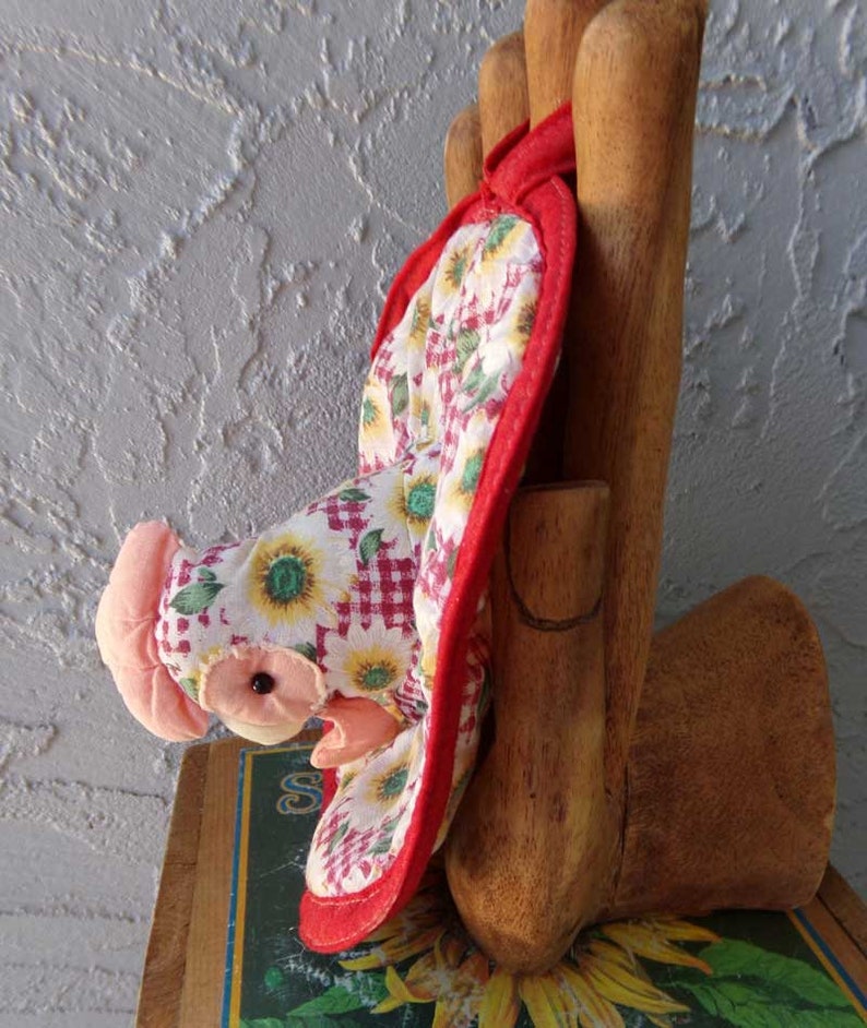 May include: Red and white quilted oven mitt shaped like a chicken with a pink head and black eyes. The mitt is hanging on a wooden hand.