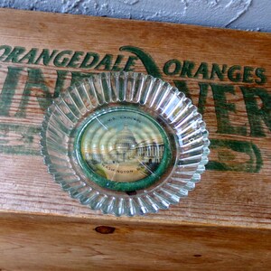 May include: A clear glass ashtray with a scalloped edge. The ashtray has a round, paper insert with a picture of the U.S. Capitol Building in Washington, D.C. The paper insert has the text "U.S. Capitol, Washington, D.C." printed on it.