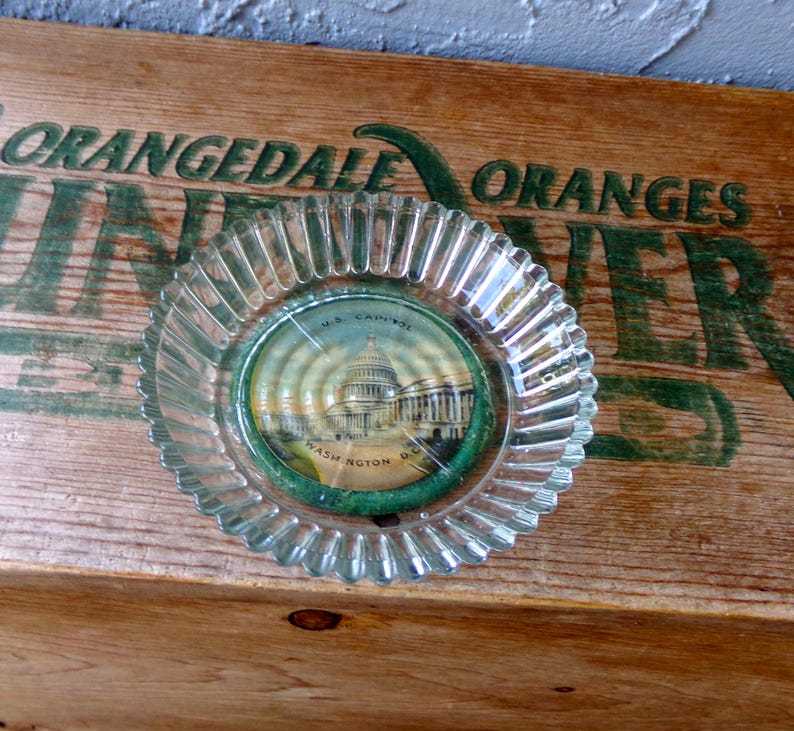 May include: A clear glass ashtray with a scalloped edge. The ashtray has a round, paper insert with a picture of the U.S. Capitol Building in Washington, D.C. The paper insert has the text "U.S. Capitol, Washington, D.C." printed on it.
