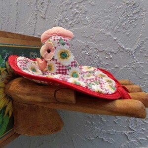 May include: A red and white gingham and sunflower print fabric chicken with a pink head and black eye, sitting on a wooden hand.