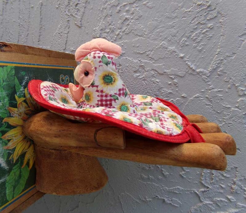 May include: A red and white gingham and sunflower print fabric chicken with a pink head and black eye, sitting on a wooden hand.