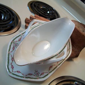 May include: A white porcelain gravy boat with a gold rim and floral design. The boat is sitting on a matching saucer with a floral design.