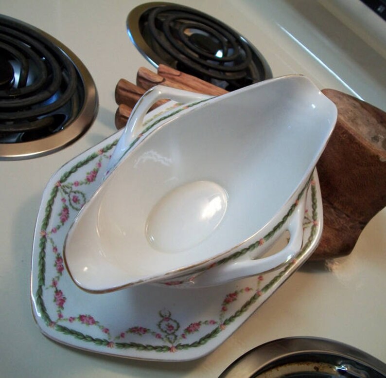May include: A white porcelain gravy boat with a gold rim and floral design. The boat is sitting on a matching saucer with a floral design.