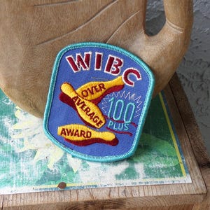 May include: A blue embroidered patch with the text "WIBC" and "OVER AVERAGE AWARD 100 PLUS". The patch features yellow and red bowling pins. The patch has a teal border.