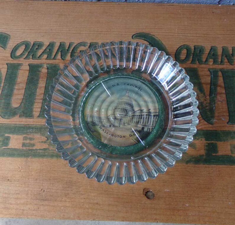 May include: A clear glass ashtray with a scalloped edge. The ashtray has a small round insert with a picture of the U.S. Capitol building in Washington D.C. The insert has the text "U.S. Capitol" and "Washington D.C."
