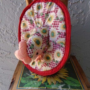 May include: A red and white pot holder with a sunflower and gingham pattern. The pot holder is shaped like a chicken with a pink beak and wattle.
