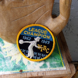 May include: A round patch with a blue and black background, featuring the text "LEAGUE CHAMPION 1962 1963" and a bowler graphic. The patch has a yellow border and the letters "WBC".