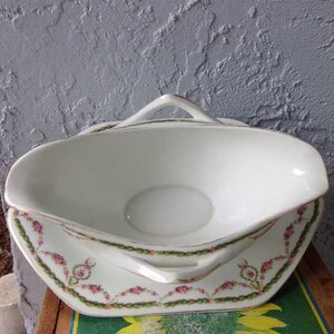May include: White porcelain gravy boat with a green and pink floral design. The boat has two handles and sits on a matching plate.
