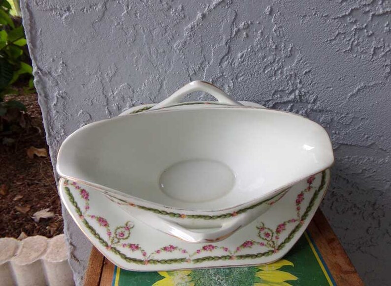 May include: White porcelain gravy boat with a green and pink floral design. The boat has two handles and sits on a matching plate.