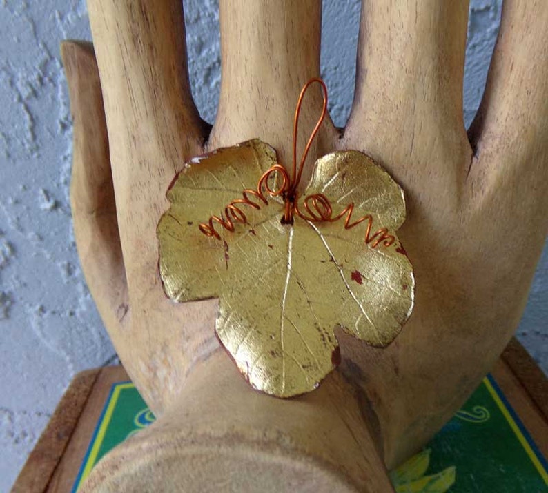 May include: A gold-colored leaf pendant with a copper wire loop and swirls. The leaf has a textured surface and is likely made of metal. The pendant is a decorative item, possibly for jewelry or crafts.
