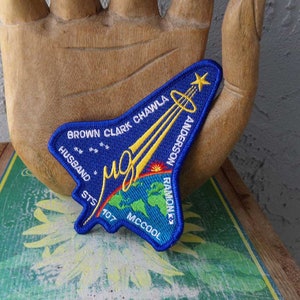 May include: A blue embroidered patch in the shape of a space shuttle. The patch features the names of the crew members, including Brown, Clark, Chawla, Anderson, Ramon, and McCool. The patch also includes the text "STS 107" and a depiction of Earth.