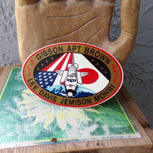 May include: A gold oval sticker with the text "GIBSON APT BROWN" and "LEE DAVIS JEMISON MOHRI" around the edge. The sticker features a space shuttle flying over a mountain range with a red, white, and blue American flag and a Japanese flag in the background.