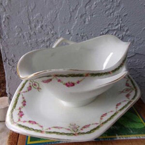 May include: A white porcelain gravy boat with a matching saucer. The boat has a gold rim and a floral design with pink roses and green leaves. The saucer has a similar design.