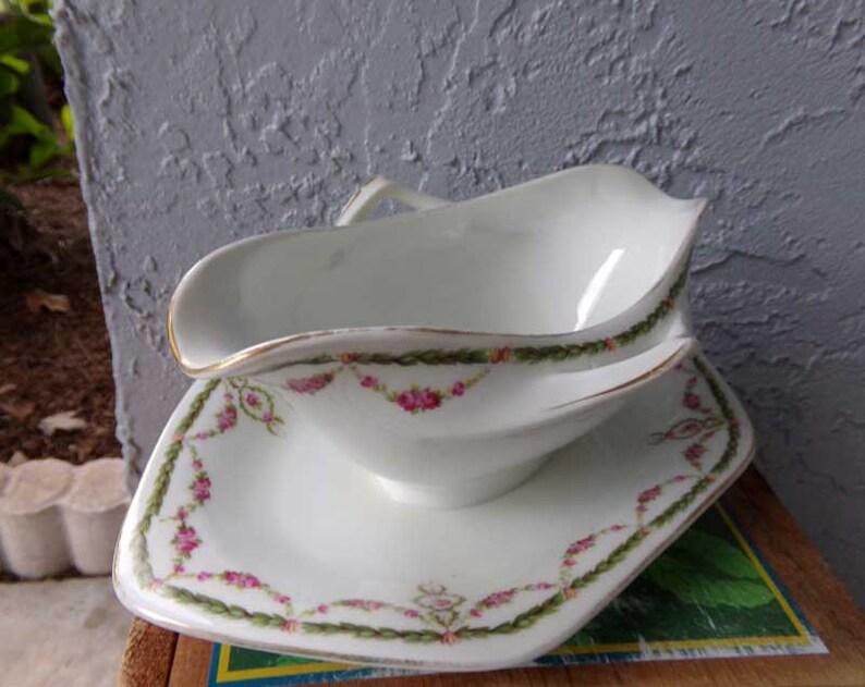 May include: A white porcelain gravy boat with a matching saucer. The boat has a gold rim and a floral design with pink roses and green leaves. The saucer has a similar design.