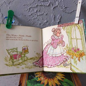 May include: An open book with an illustration of a woman in a pink dress and a white apron. The text on the page reads "Old Mother Middle Muddle put down her knitting and went to the kitchen to brew a cup of tea."