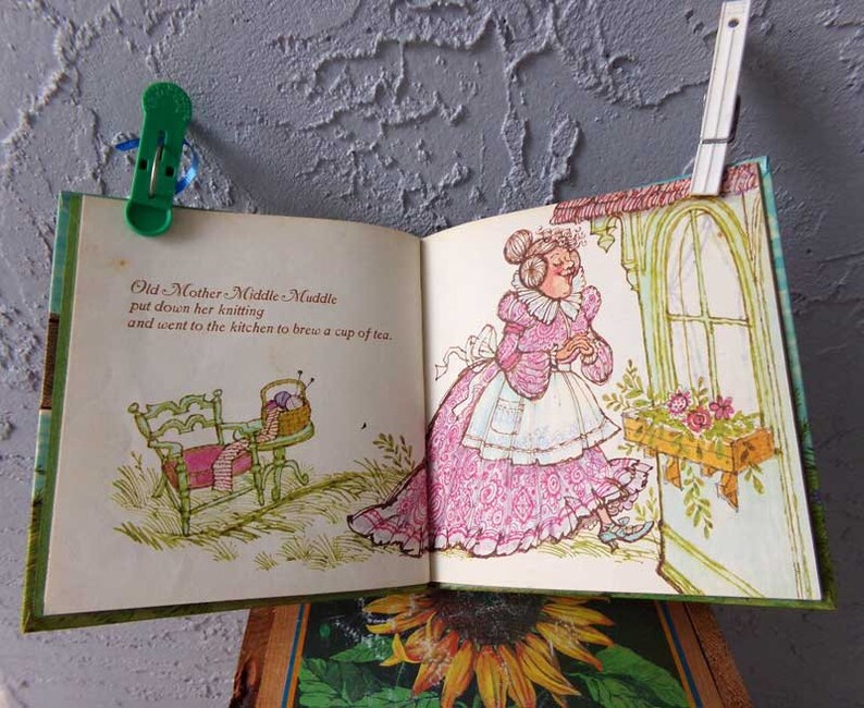 May include: An open book with an illustration of a woman in a pink dress and a white apron. The text on the page reads "Old Mother Middle Muddle put down her knitting and went to the kitchen to brew a cup of tea."